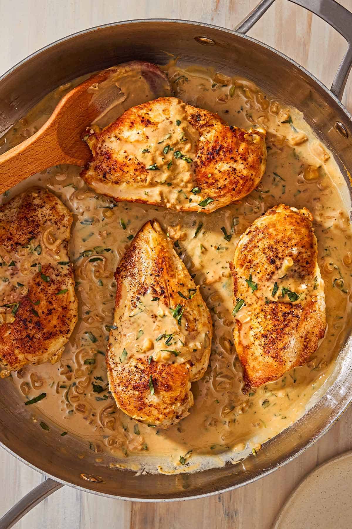 An overhead photo of 4 chicken breast halves and the tarragon mustard sauce in a skillet with a wooden spoon.