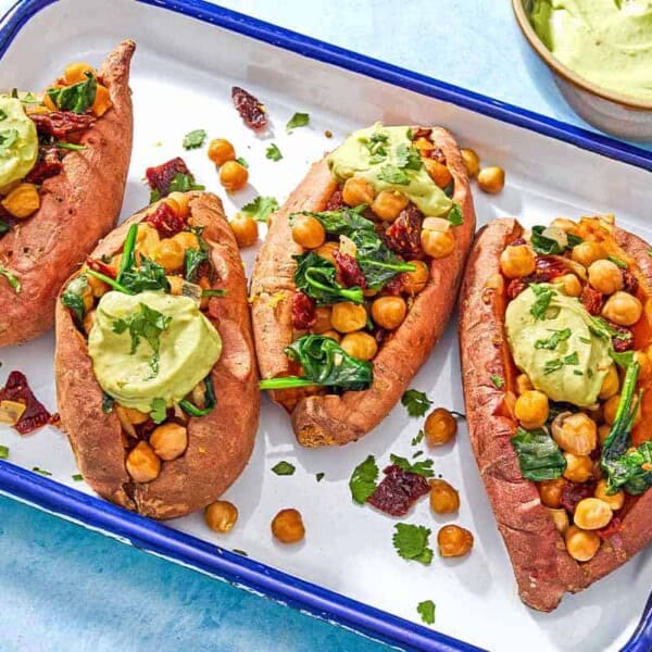 A close up of 4 stuffed sweet potatoes with avocado crema on a platter next to a bowl of avocado crema.