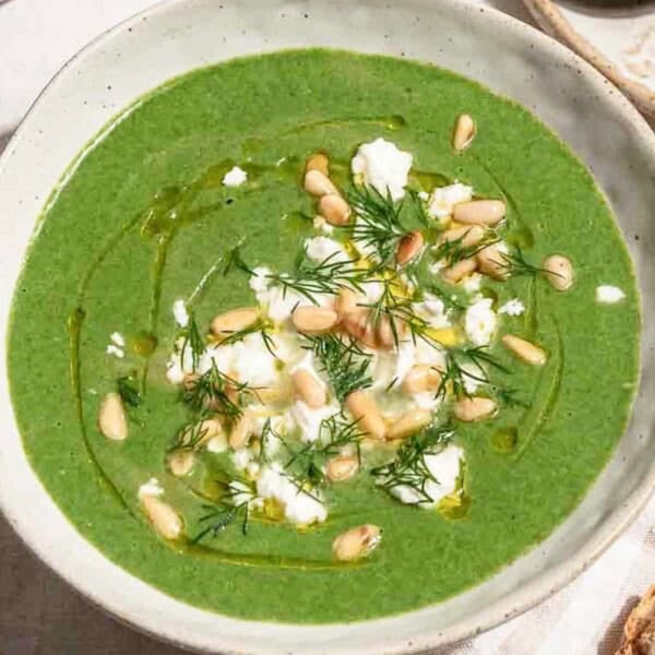 A close up of a serving of spinach soup in a bowl topped with dill, pine nuts, crumbled feta and a drizzle of olive oil. Next to this is a bowl of salt and pepper.
