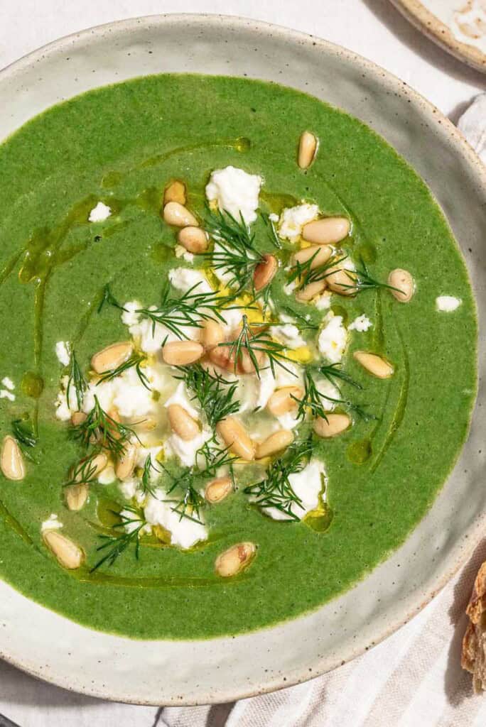 A close up of a serving of spinach soup in a bowl topped with dill, pine nuts, crumbled feta and a drizzle of olive oil. Next to this is a sprig of dill and a small bowl of pine nuts.
