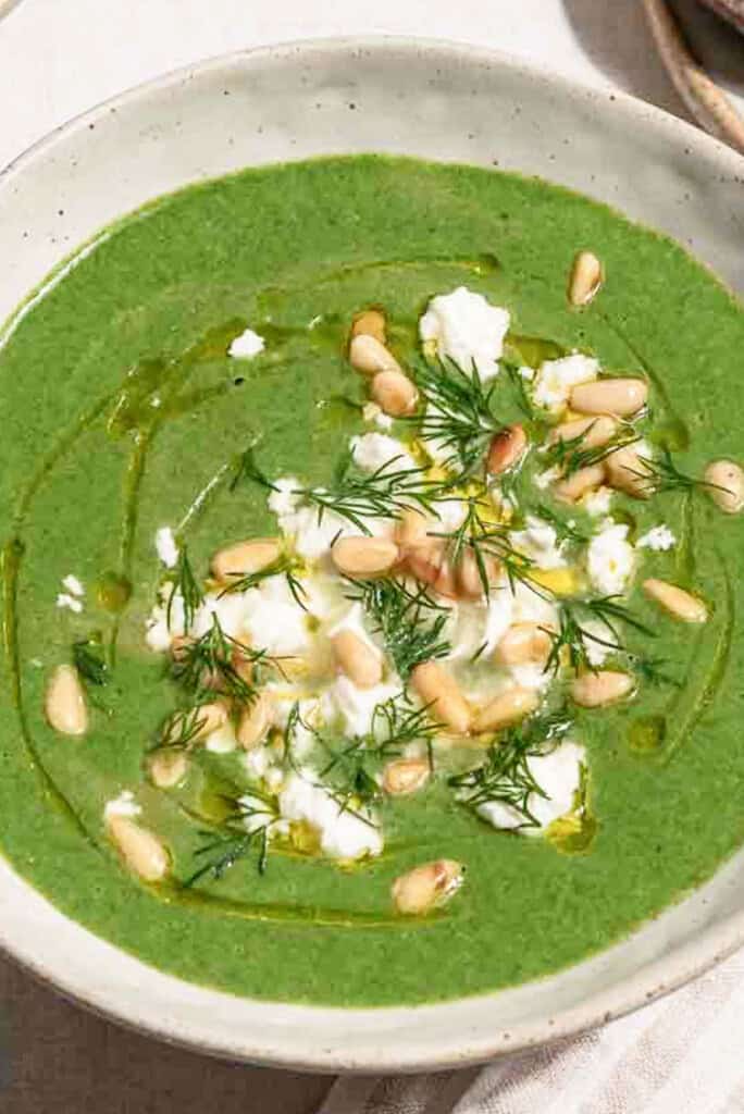 A close up of a serving of spinach soup in a bowl topped with dill, pine nuts, crumbled feta and a drizzle of olive oil.