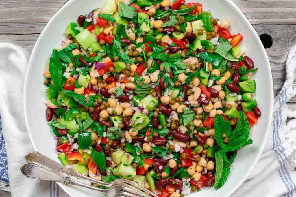 An overhead photo of a Mediterranean three bean salad in a serving bowl with two forks. Next to this is a kitchen towel.