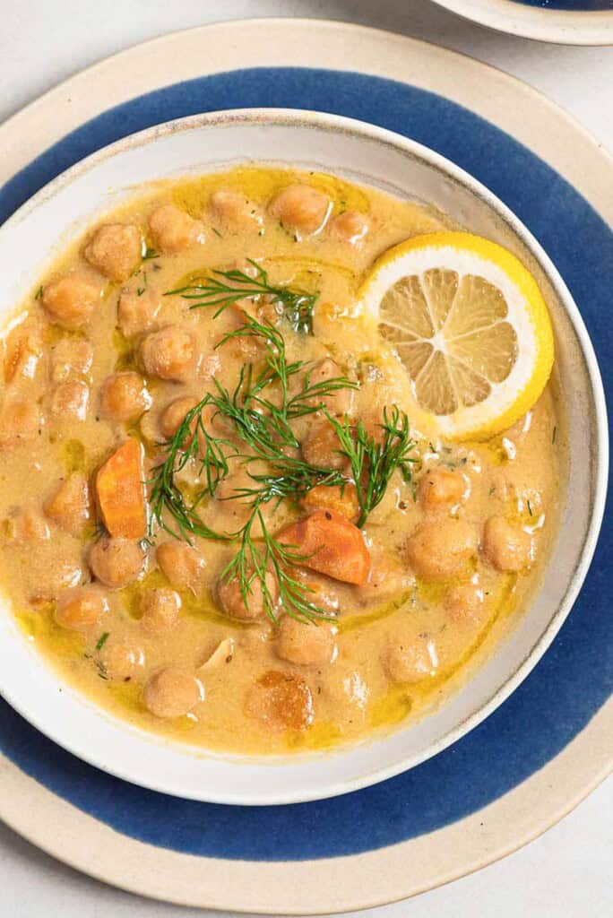 A close up of revithosoupa Greek chickpea soup garnished with dill, a drizzle of olive oil and a lemon slice in a bowl resting on a plate.