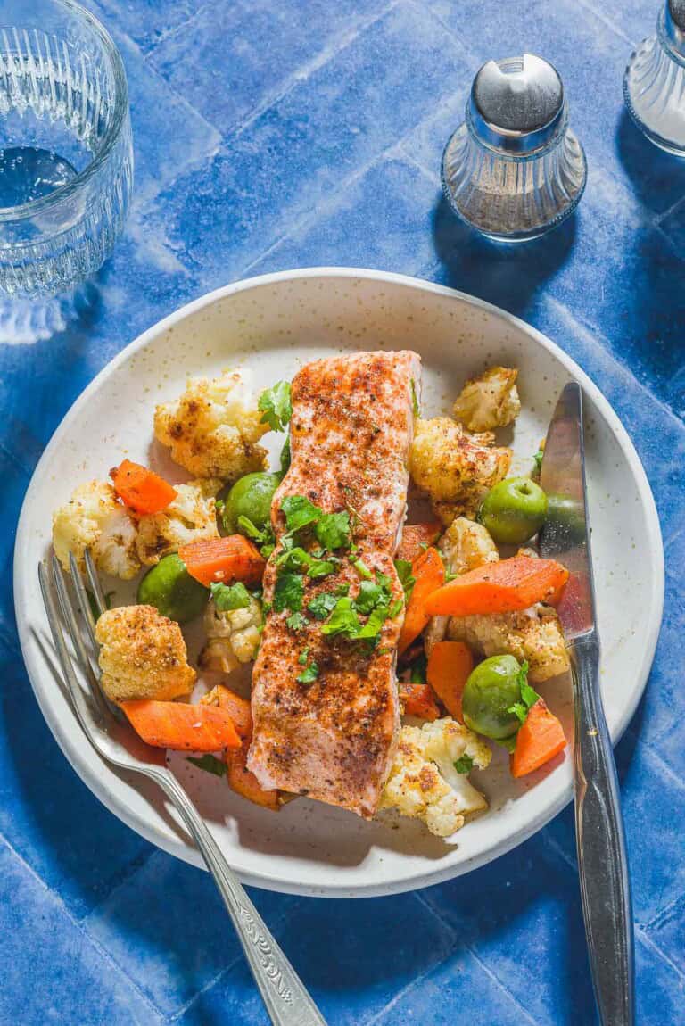 An overhead photo of a baharat roasted salmon fillet on a bed of vegetables and olives on a plate with a fork and knife. Next to this is a glass of water and salt and pepper shakers.