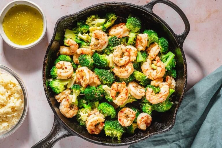 An overhead photo of the cooked shrimp and broccoli skillet. Next to this is a cloth napkin, a bowl of couscous, and a bowl of ladolemono sauce.