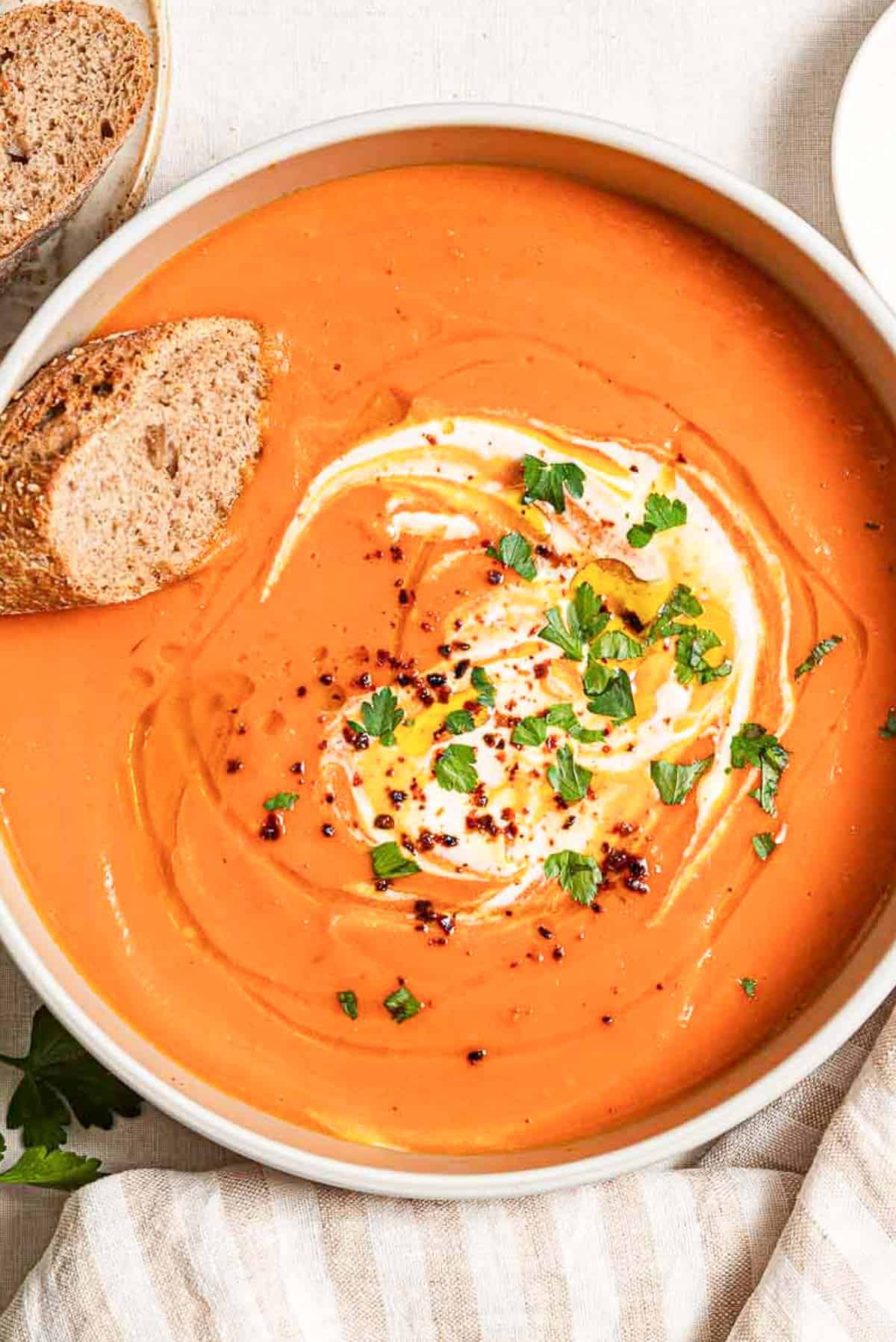An overhead photo of sweet potato soup topped with a dollop of greek yogurt, a drizzle of olive oil, Aleppo pepper and chopped parsley in a bowl along with a slice of bread. Next to this is a cloth napkin.