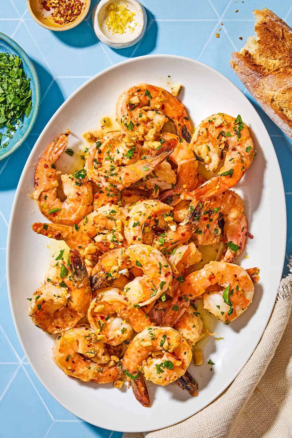 An overhead photo of shrimp scampi on a platter. Next to this is a cloth napkin, a loaf of crusty bread, and bowls of lemon zest, red pepper flakes and chopped parsley.