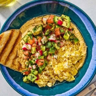 A protein packed breakfast hummus bowl with a pita wedge next to a fork, a spoon and a bowl of za'atar.