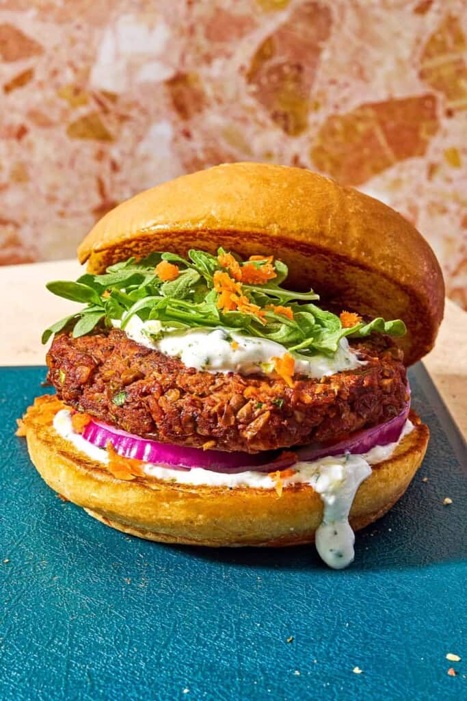 A vegetarian burger on a bun with yogurt herb sauce, red onion, grated carrots and arugula leaves.