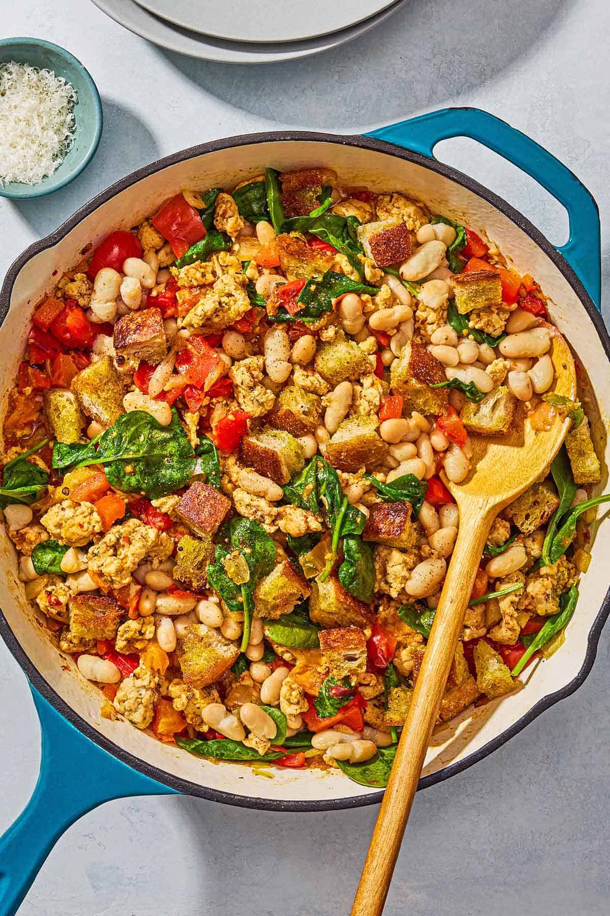 The ground turkey dinner in a skillet with a wooden spoon next to a stack of two plates and a small bowl of grated parmesan cheese.