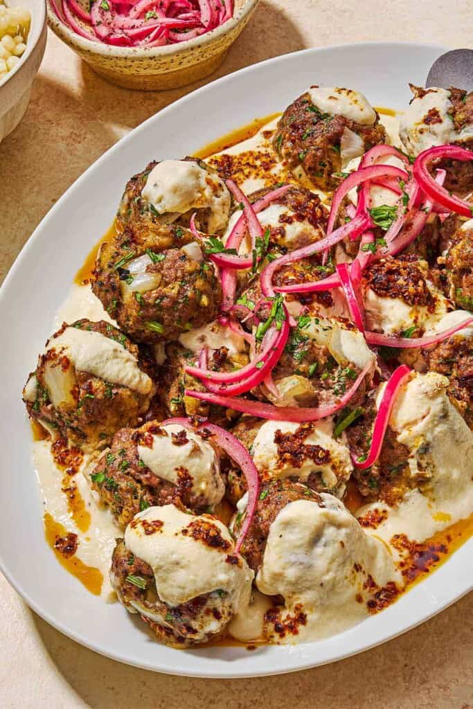 The turkish meatballs and yogurt sauce topped with pickled red onions and chopped parsley on a platter next to a bowl pickled red onions.