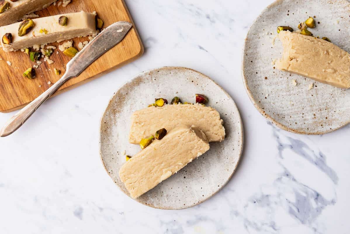 Slices of halva topped with roasted pistachios on 2 plates next to the cutting board with the rest of the halva and a knife.
