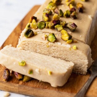 A loaf of halvah topped with roasted pistachios with 2 slices cut on a cutting board with a knife.