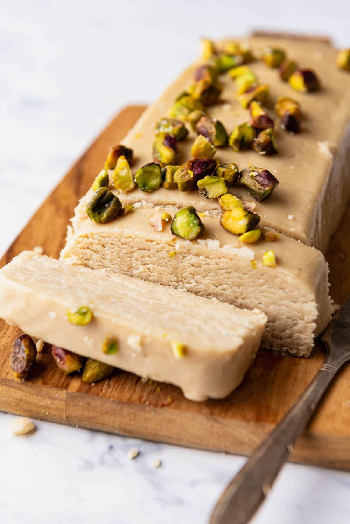 A loaf of halvah topped with roasted pistachios with 2 slices cut on a cutting board with a knife.