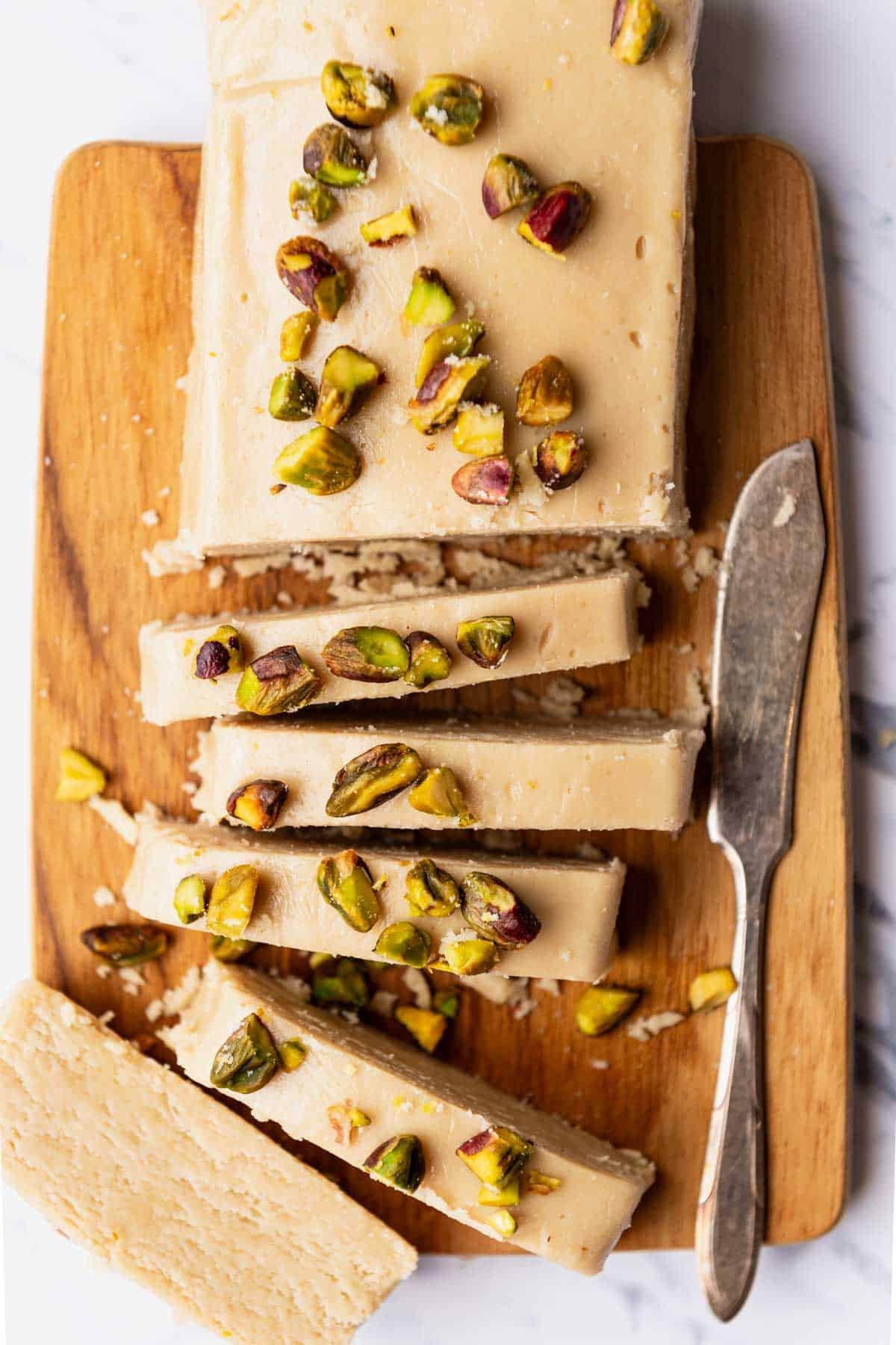 A loaf of halvah topped with roasted pistachios with several slices cut on a cutting board with a knife.