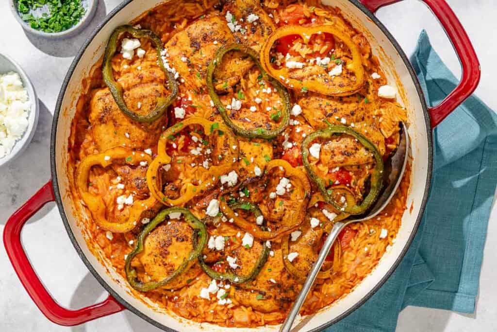 The one pan chicken orzo in a skillet with a serving spoon next to bowls of crumbled feta and chopped parsley, and a cloth napkin.