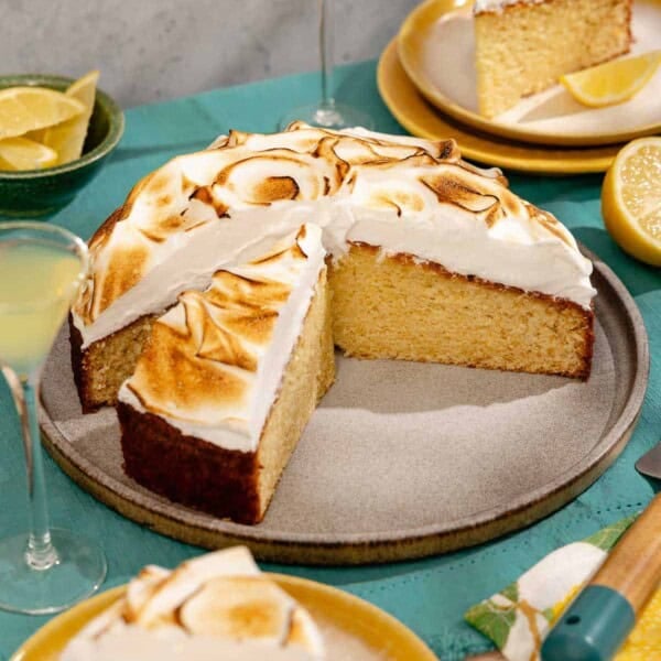 A limoncello cake on a platter with a piece cut from it. Next to this is a bowl of lemon wedges, two plates each with a piece of the cake, 2 glasses of limoncello, and a lemon half.
