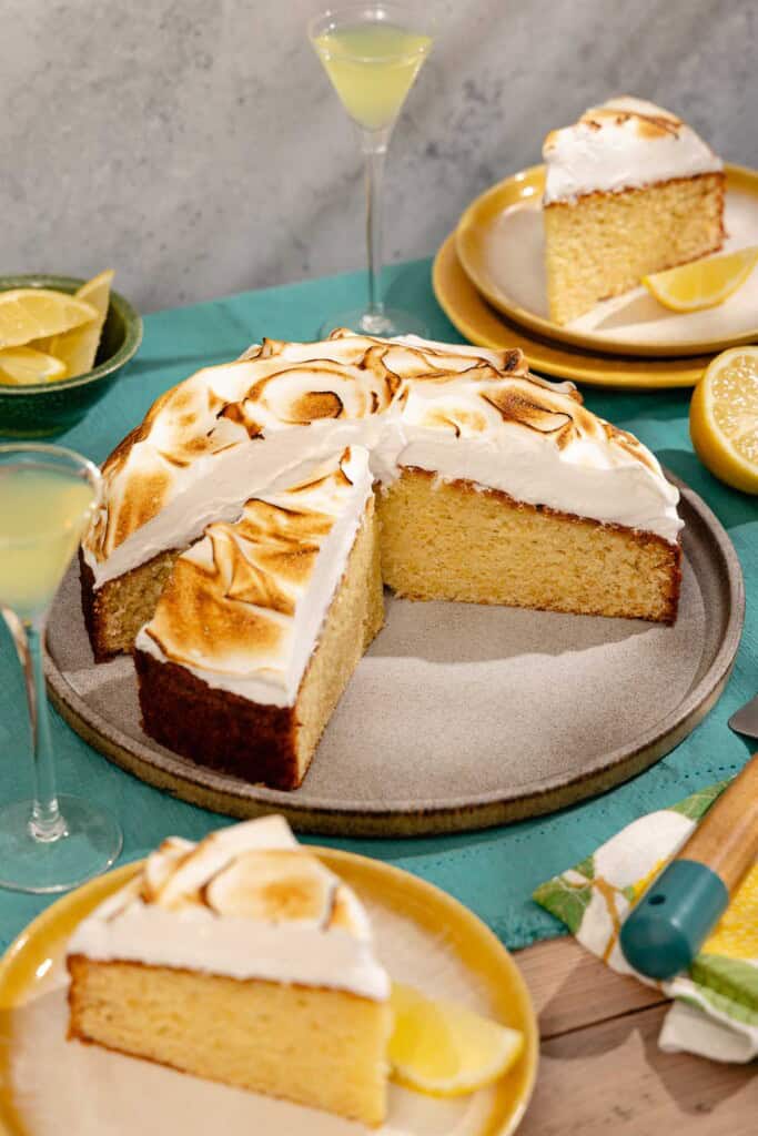 A limoncello cake on a platter with a piece cut from it. Next to this is a bowl of lemon wedges, two plates each with a piece of the cake, 2 glasses of limoncello, and a lemon half.