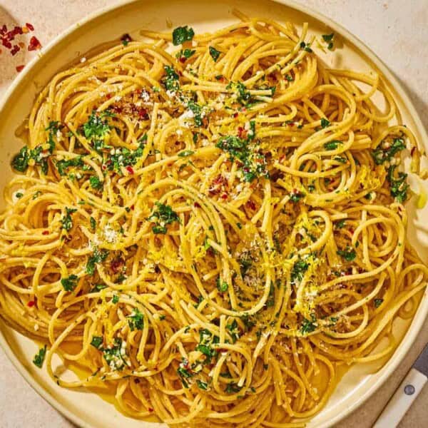 Lemon pasta topped with parsley, lemon zest, red pepper flakes and parmesan on a plate next to a fork and small bowls of red pepper flakes and parmesan cheese.