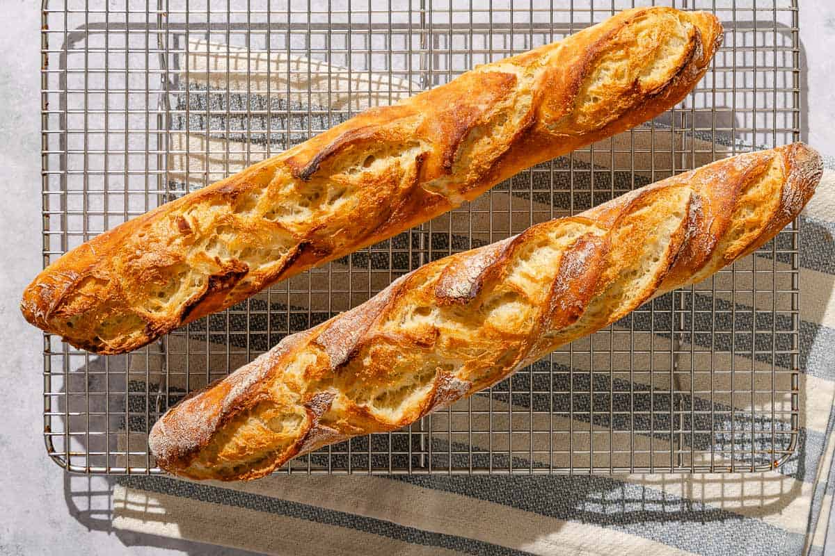 Two baked French baguettes cooling on a wire rack.