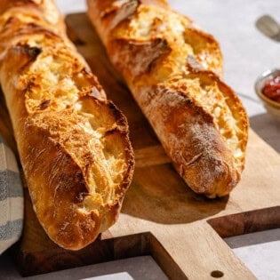 Two baked french baguettes on a wooden serving board next to a cloth kitchen towel and a bowl of jam with a spoon.