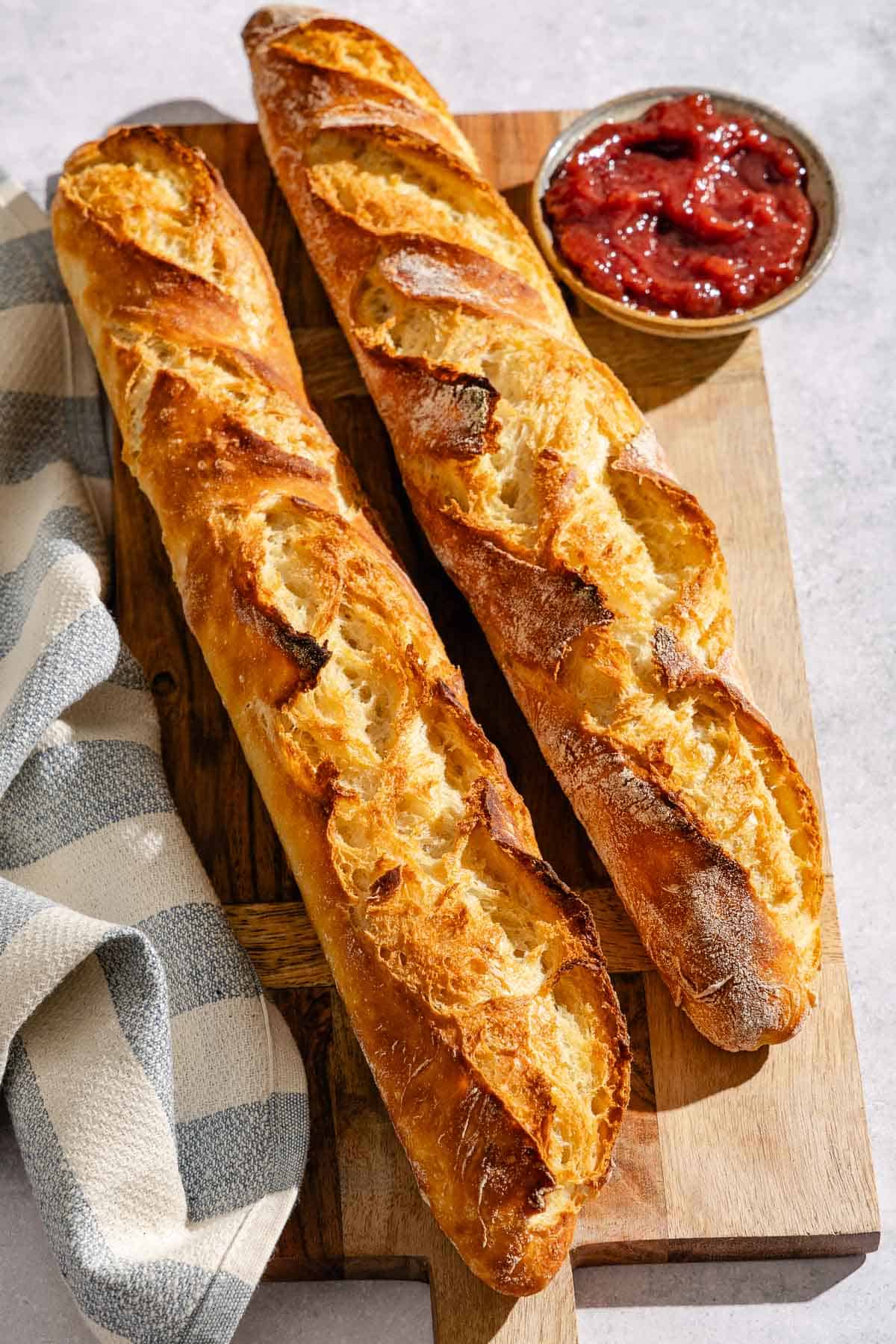 2 baked baguettes on a wooden serving board next to a bowl of jam and a cloth kitchen towel.