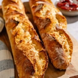 Two baked baguettes on a wooden serving board. Next to this is a cloth kitchen towel and a bowl of jam with a spoon.