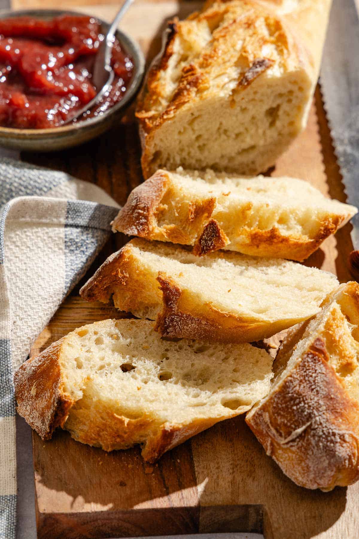 A sliced baguette on a wooden serving board along with a knife and a bowl of jam.
