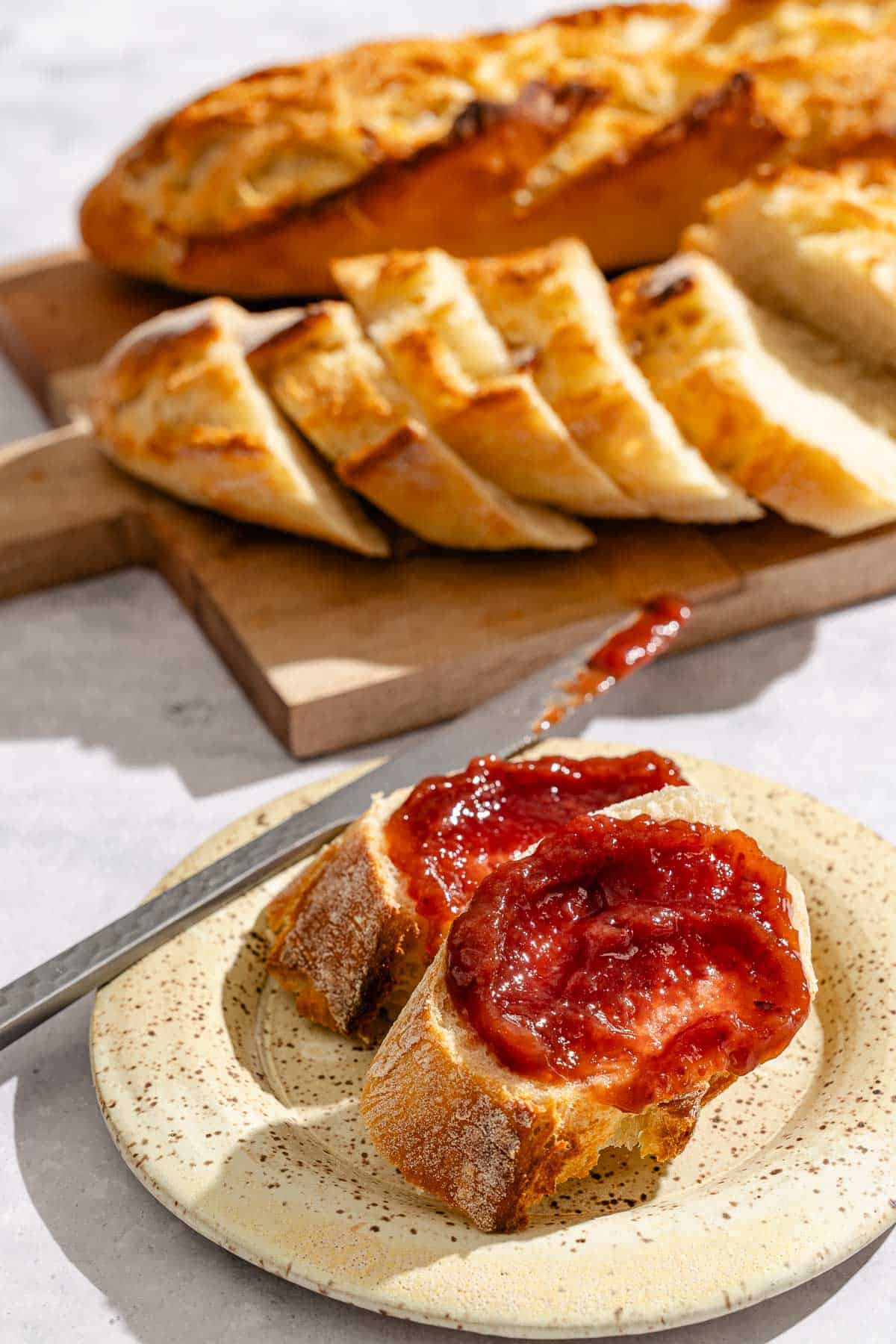 A close up of 2 slices of the baguette topped with jam on a plate with a knife. In the background is a wooden serving board with a whole baguette and a sliced baguette.