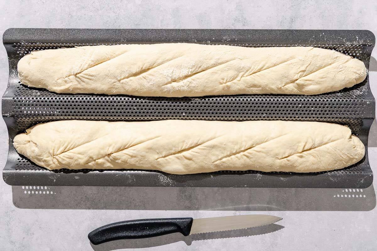 Two unbaked scored French baguettes on a wire rack next to a sharp knife.