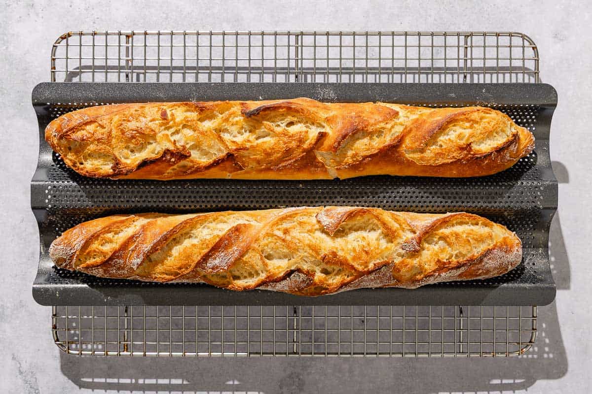 Two baked French baguettes cooling on a wire rack.