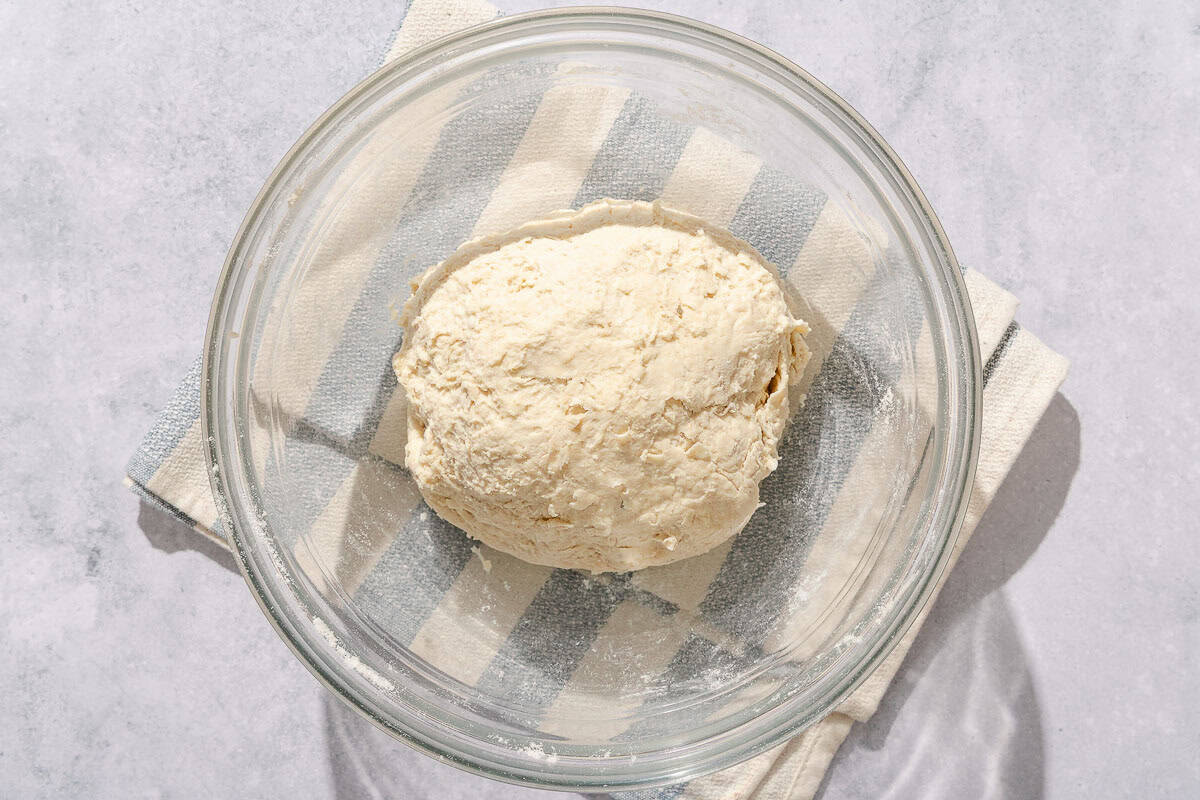 A ball of dough in a glass mixing bowl resting on a cloth kitchen towel.