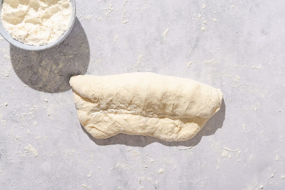 The dough for the baguette being rolled out onto a countertop dusted in flour. Next to this is a small bowl of flour.