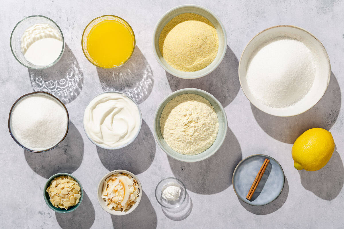 Ingredients for basbousa including sugar, greek yogurt, fine semolina, coarse semolina, baking powder, milk, butter, shredded coconut, sliced almonds, a cinnamon stick and a lemon.