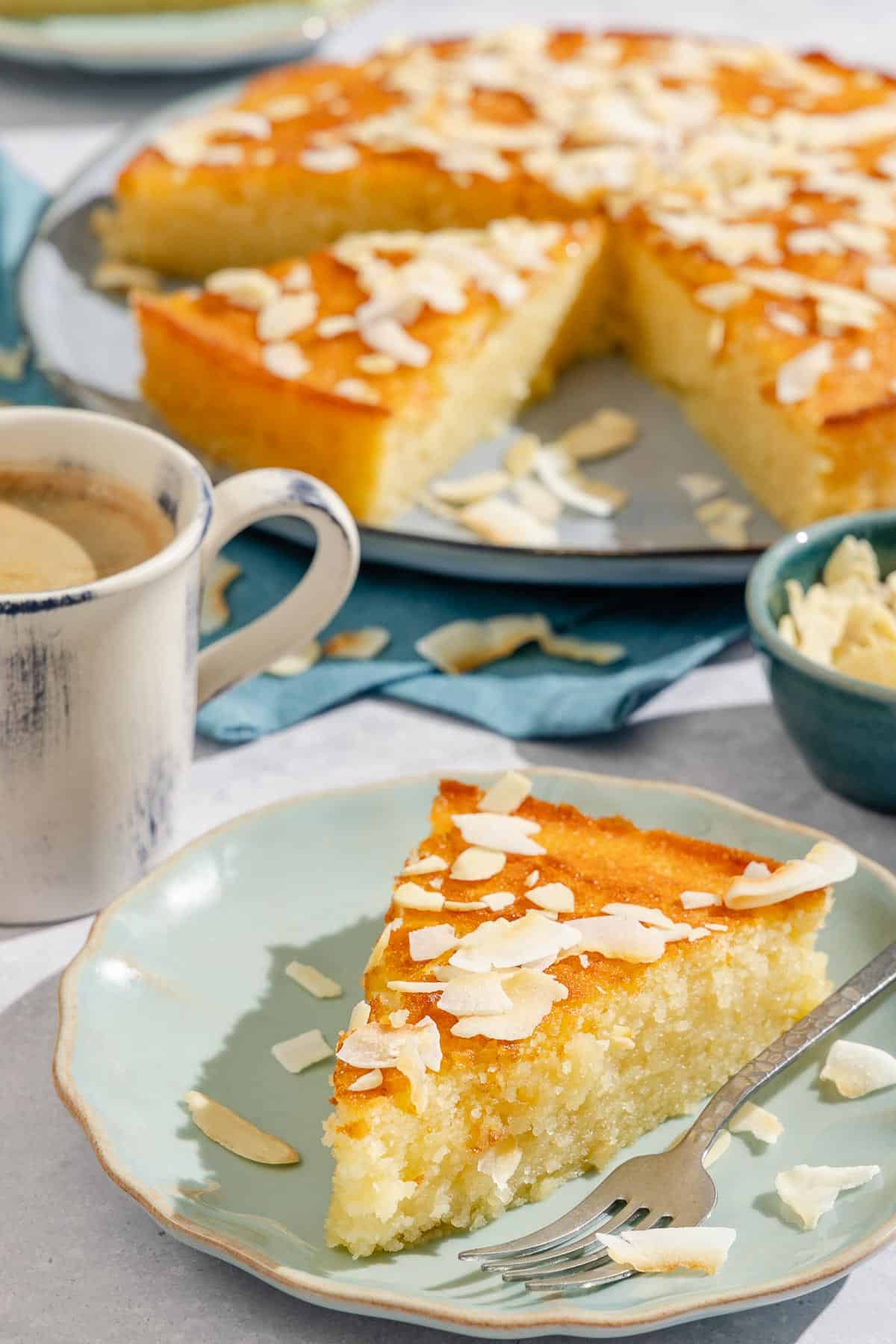A slice of basbousa on a plate with a fork next to a cup of coffee, a bowl of sliced almonds, and the rest of the cake on a platter.