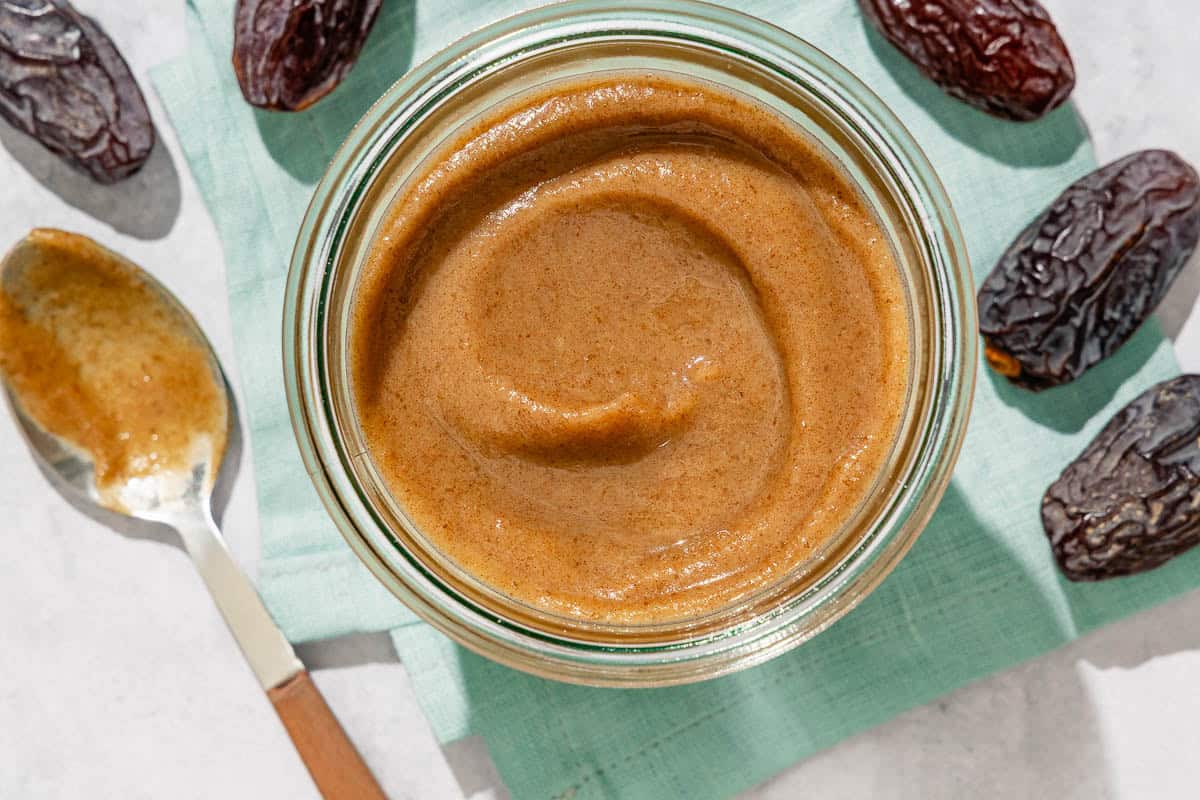 Date Caramel in a jar sitting on a cloth napkin surrounded by some dates and a spoon.
