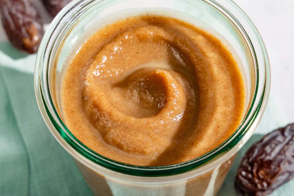Date Caramel in a jar sitting on a cloth napkin surrounded by some dates and a spoon.