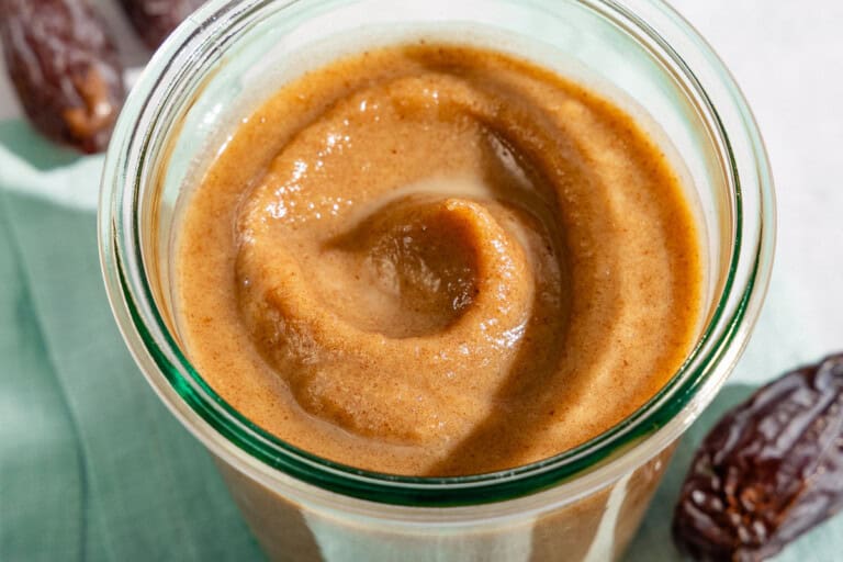 Date Caramel in a jar sitting on a cloth napkin surrounded by some dates and a spoon.