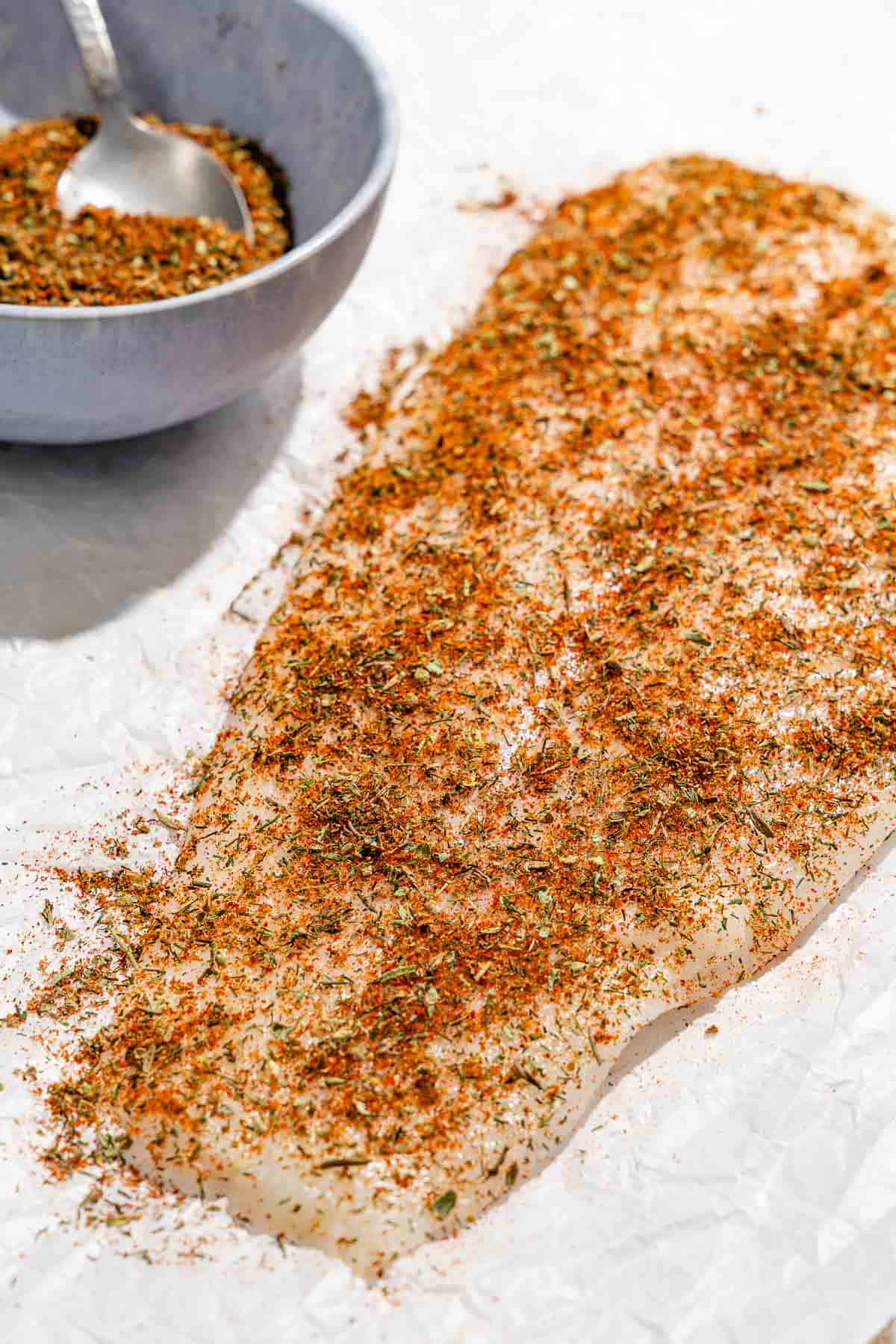 A white fish fillet coated with the fish seasoning next to a bowl with the rest of the seasoning and a spoon.