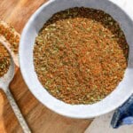 Fish seasoning in a bowl on a wooden tray next to a spoon with some of the seasoning and a cloth kitchen towel.