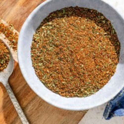 Fish seasoning in a bowl on a wooden tray next to a spoon with some of the seasoning and a cloth kitchen towel.
