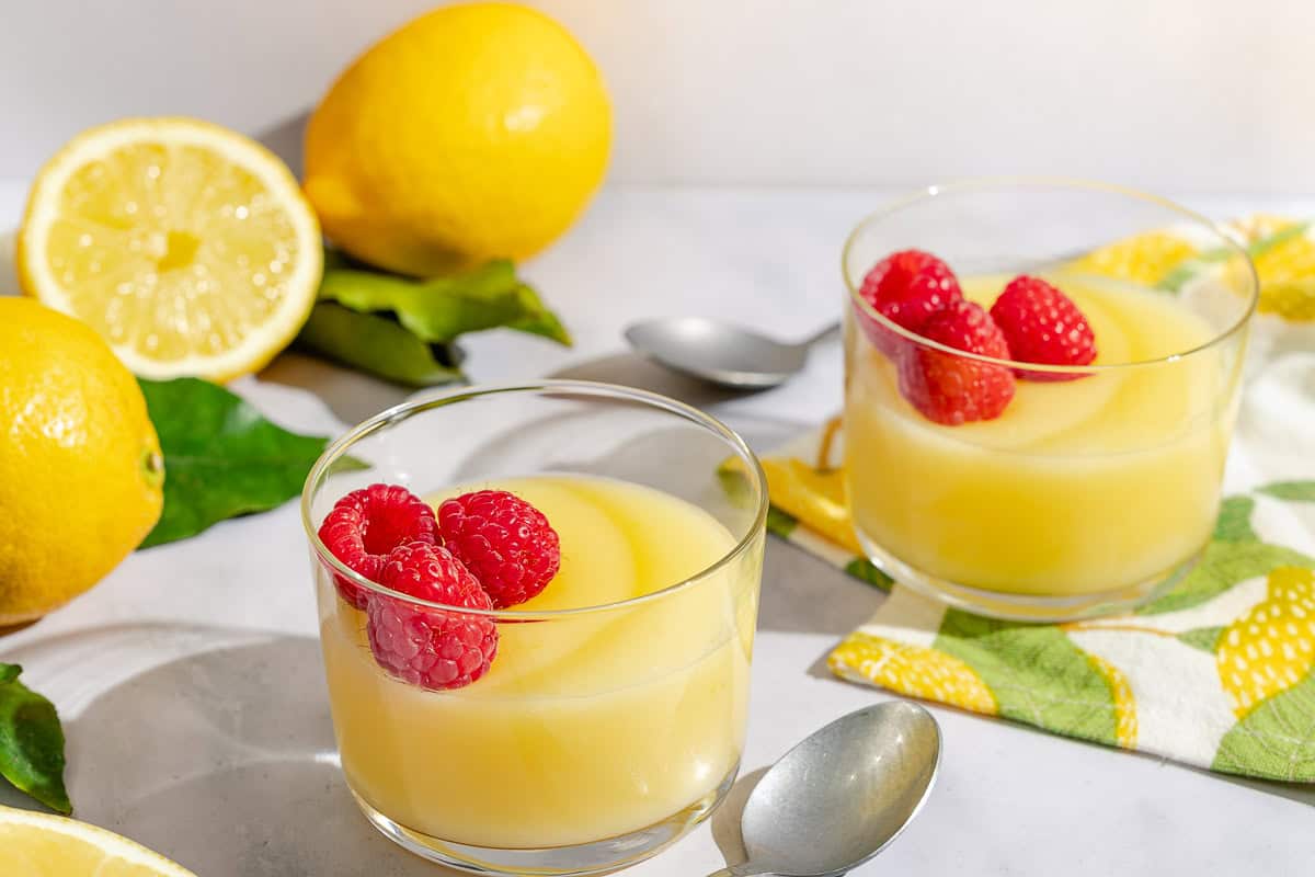 Lemon puddo seasoned with raspberries in a small glass bowl next to a spoon, another bowl of pudding and several lemons.