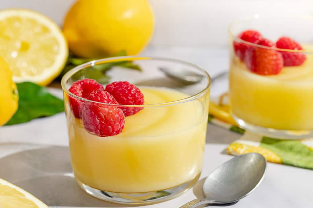Lemon pudding topped with raspberries in a small glass bowl next to a spoon, another bowl of pudding, and several lemons.