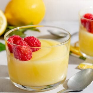 Lemon pudding topped with raspberries in a small glass bowl next to a spoon, another bowl of pudding, and several lemons.