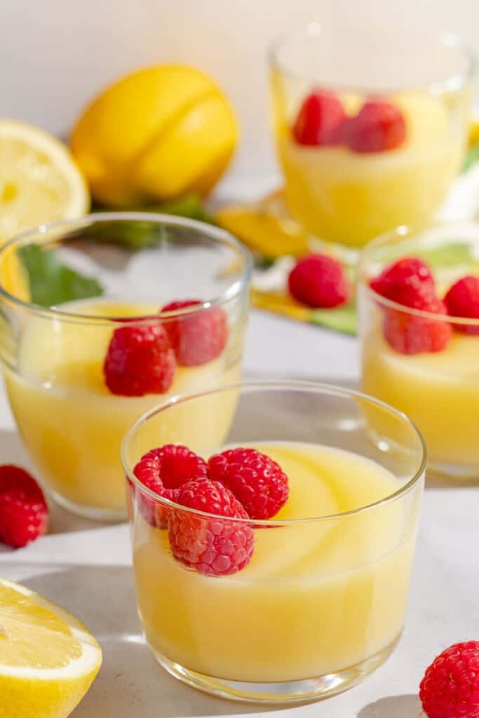 Four small glass bowls of lemon pudding, each topped with raspberries. Next to these are several lemons.