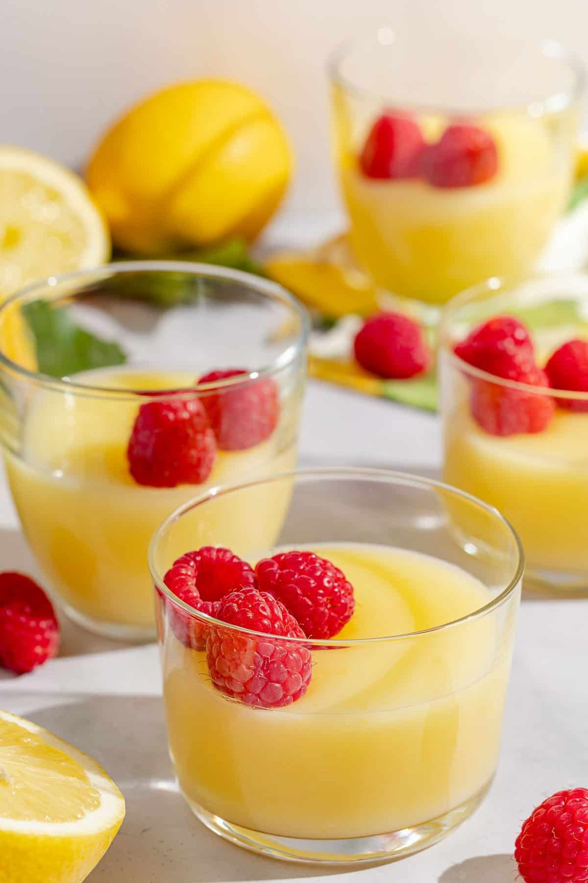 Four small lemon pudding glass bowls, each seasoned with raspberries. Next to these there are several lemons.