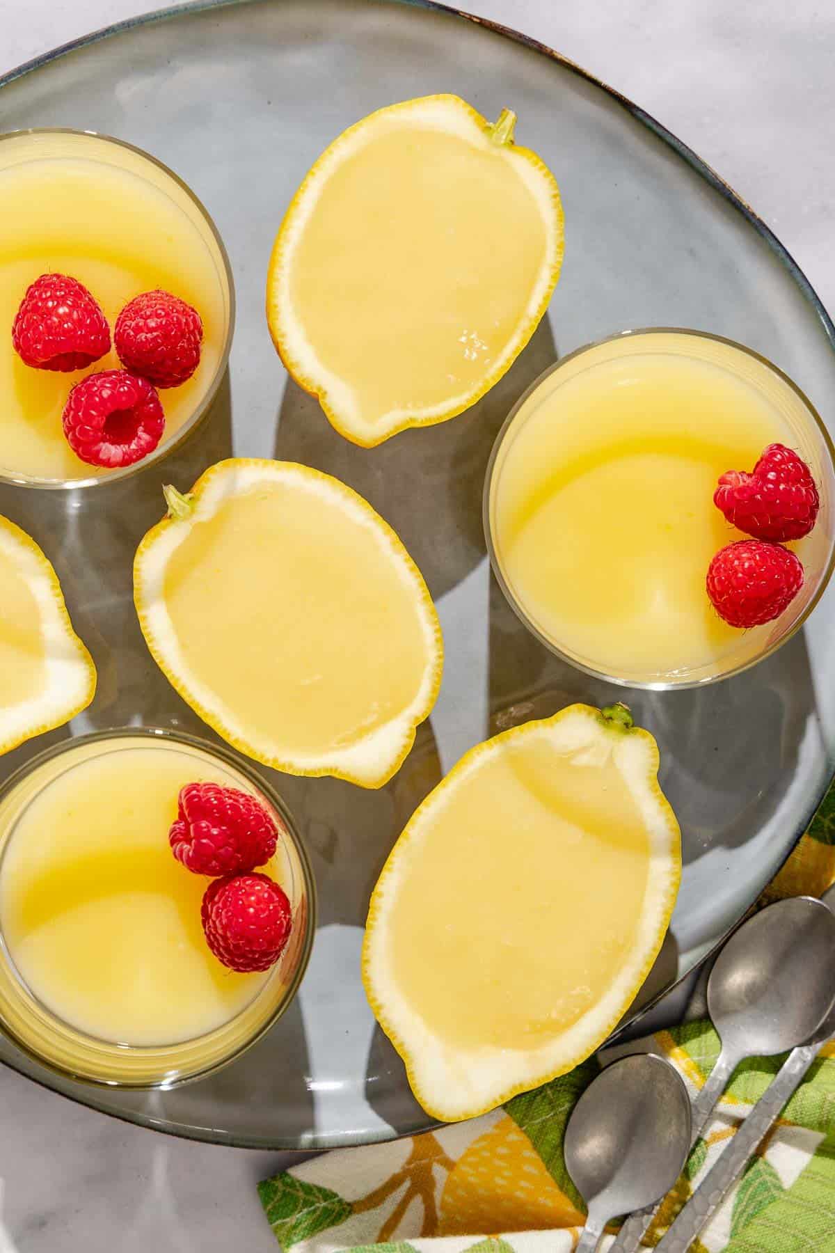 Three small glass bowls and 4 half of the lemon fill with the lemon pudding on a plate. Next to this there is a fabric napkin and 3 tablespoons.
