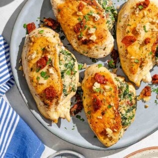 Four spinach stuffed chicken breasts on a platter surrounded by a cloth napkin, bowls of chopped walnuts and parsley, and a stack of 2 plates.