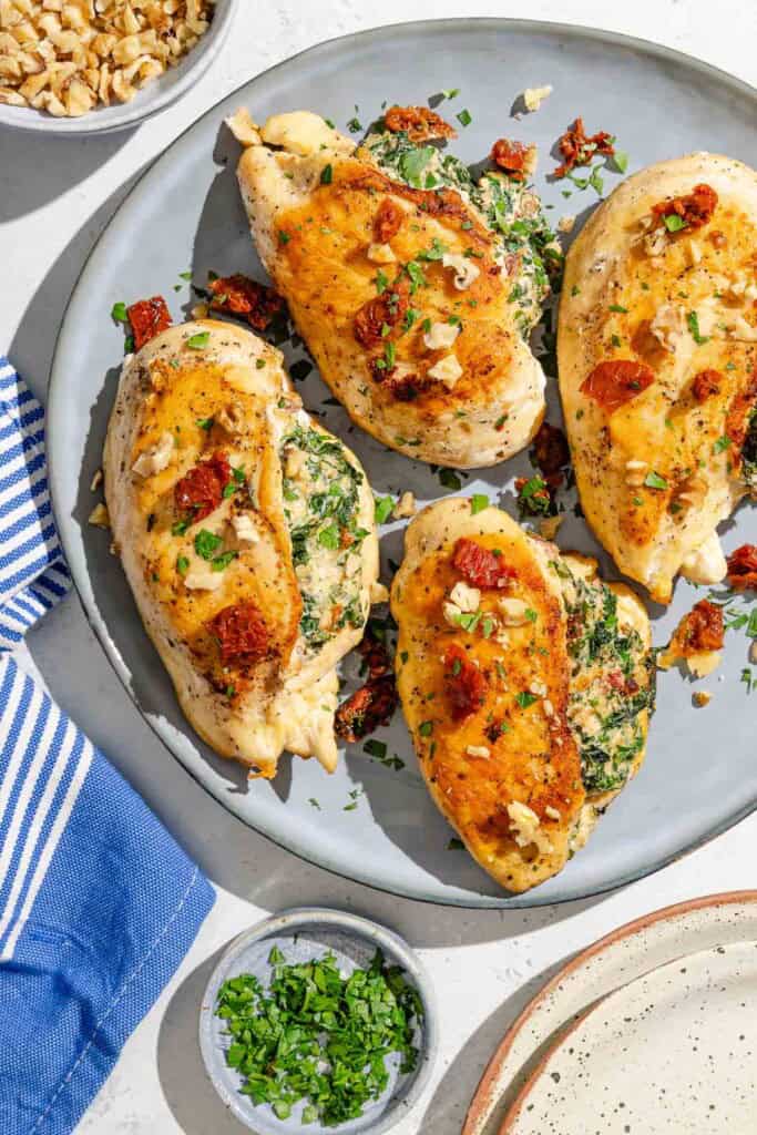 Four spinach stuffed chicken breasts on a platter surrounded by a cloth napkin, bowls of chopped walnuts and parsley, and a stack of 2 plates.