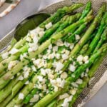Steamed asparagus topped with a vinaigrette, grated eggs and minced chives on a platter with a serving spoon. Next to this is a cloth napkin, a stack of 2 plates, and a bowl of minced chives.